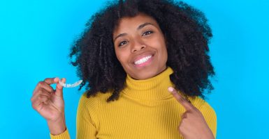 MODEL holding an invisible aligner and pointing to her perfect straight teeth. Dental healthcare and confidence concept.