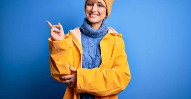 Young blonde woman with short hair wearing rain coat for rainy weather over blue background with a big smile on face, pointing with hand and finger to the side looking at the camera.