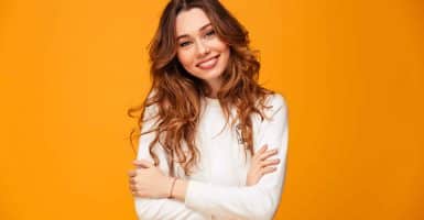 Smiling brunette woman in sweater posing with crossed arms and looking at the camera over yellow background